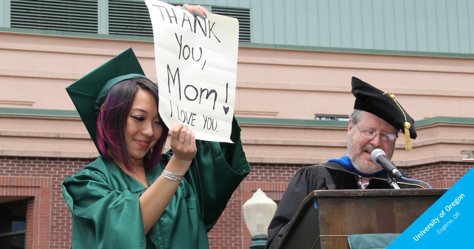 Asian female graduate holding a sign that says "Thank you, Mom. I love you."